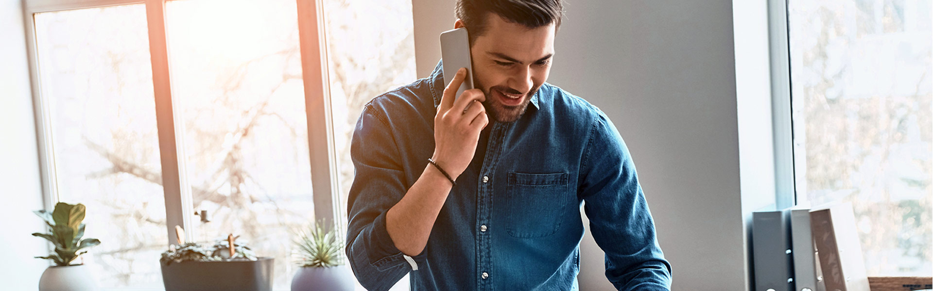 Young man standing at his desk smiling while talking on the phone and looking at his computer