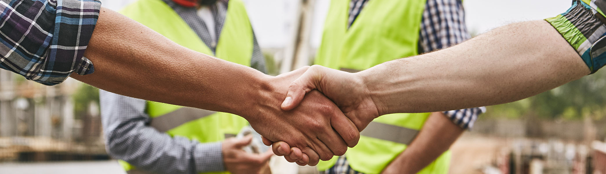 We did it! Close up photo of builders shaking hands against cheerful colleagues while working together at construction site. Team work.