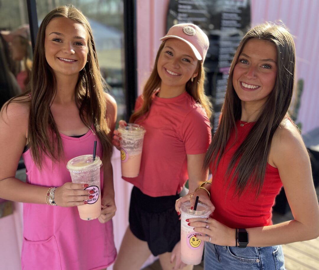 Three teenage girls holding drinks standing outside a dirty soda stand